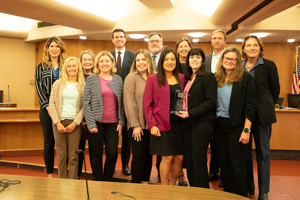 Group of 12 people, standing in the Board of Supervisors chambers, one person in the middle holding an award.