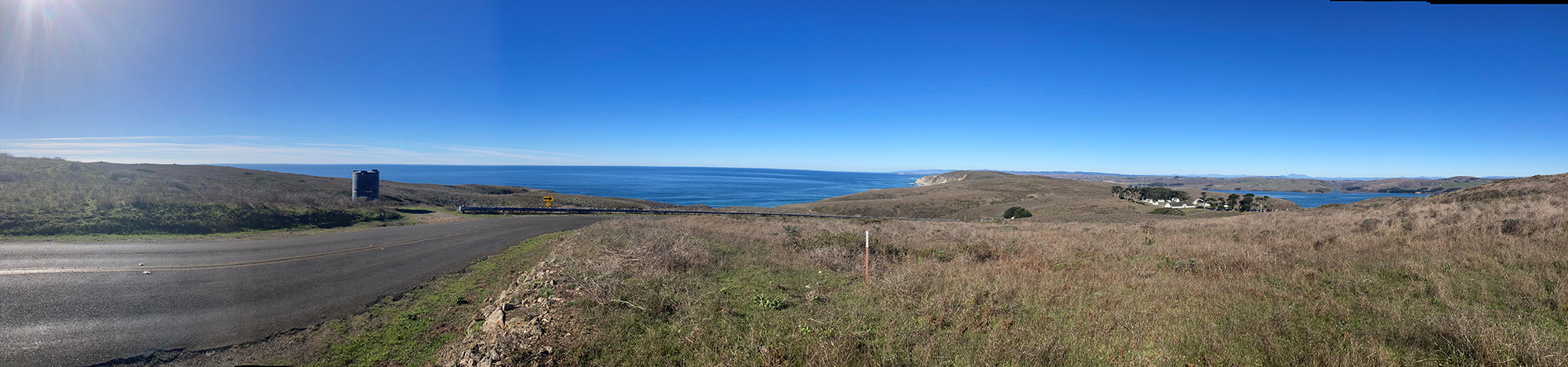 Road with green and brown grass on the sides and the ocean in front.