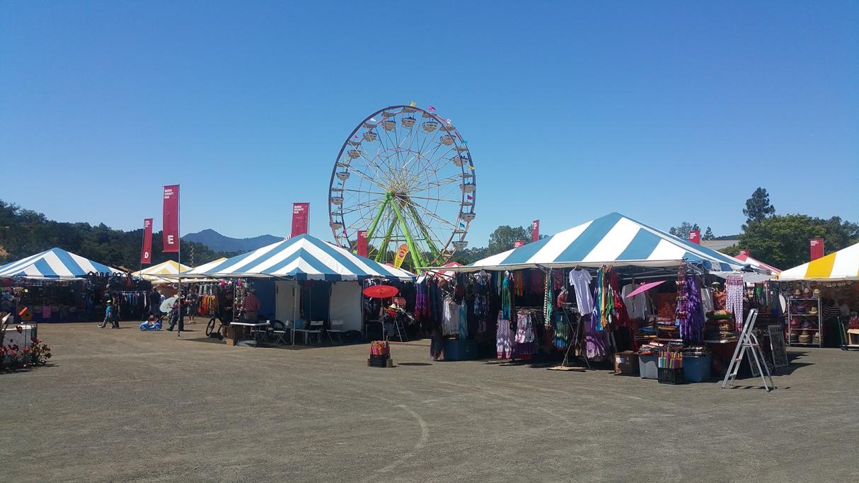 A scene of the Marin County Fairgrounds with vendor tents and people in the foreground and the ferriswheel in the background.