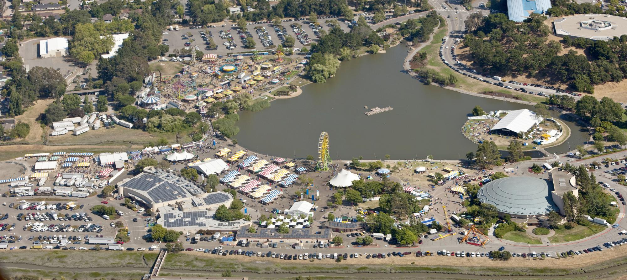 Aerial, panoramic view of the Fairgrounds showing the parking area, Lagoon Park with its island, the layout of the vendor and exhibit tents, the exhibit hall and other Civic Center buildings.