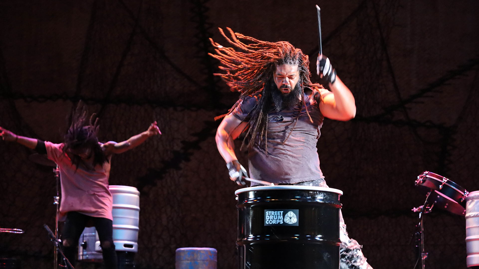 A man plays the steel drum, while another man dances behind him. 