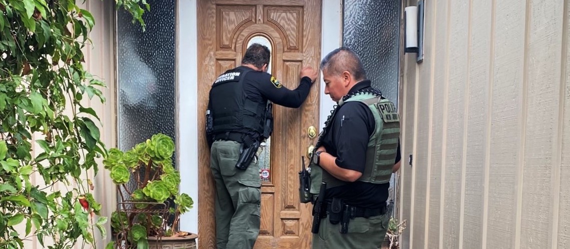 Two male law enforcement officers stand outside an offender's front door, as one of them knocks.