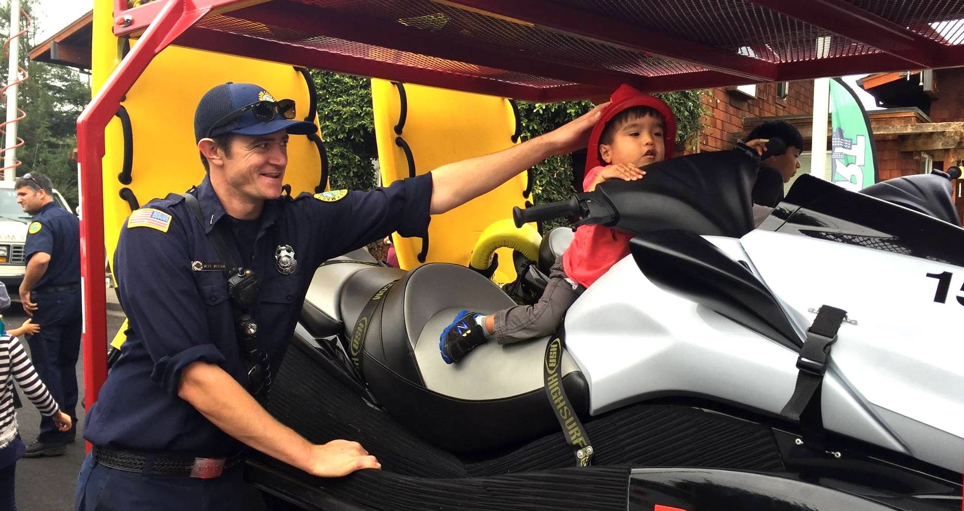 A smiling male firefighter gently holds the head of a young boy who is setting on the fire department's personal watercraft at a community event.