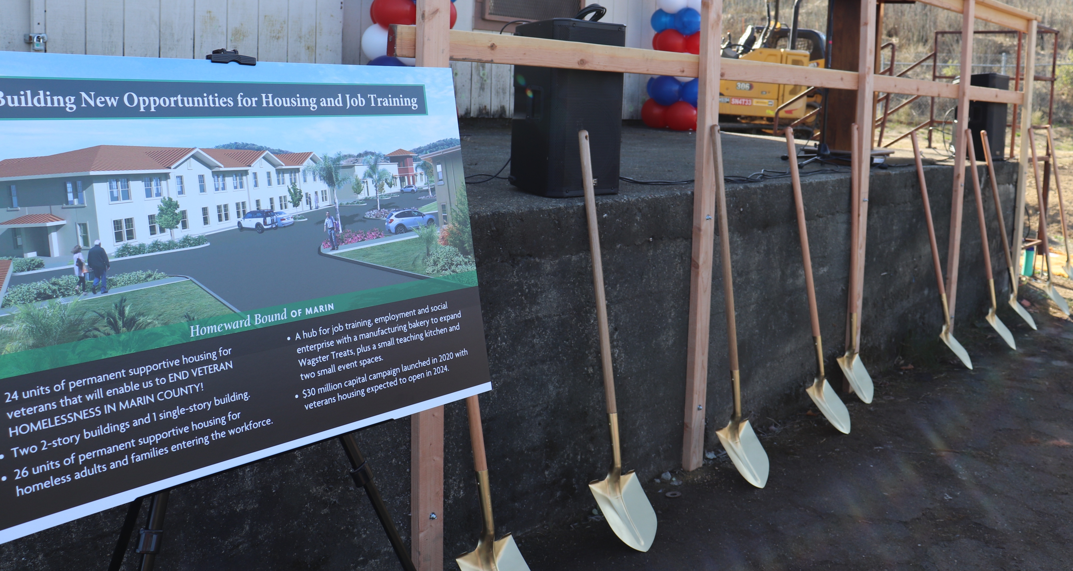 A view of a sign of 4 feet by 3 feet showing an architectural rendering of the new housing proposed for Novato. A line of golden shovels sits next to the sign to be used for a groundbreaking ceremony.