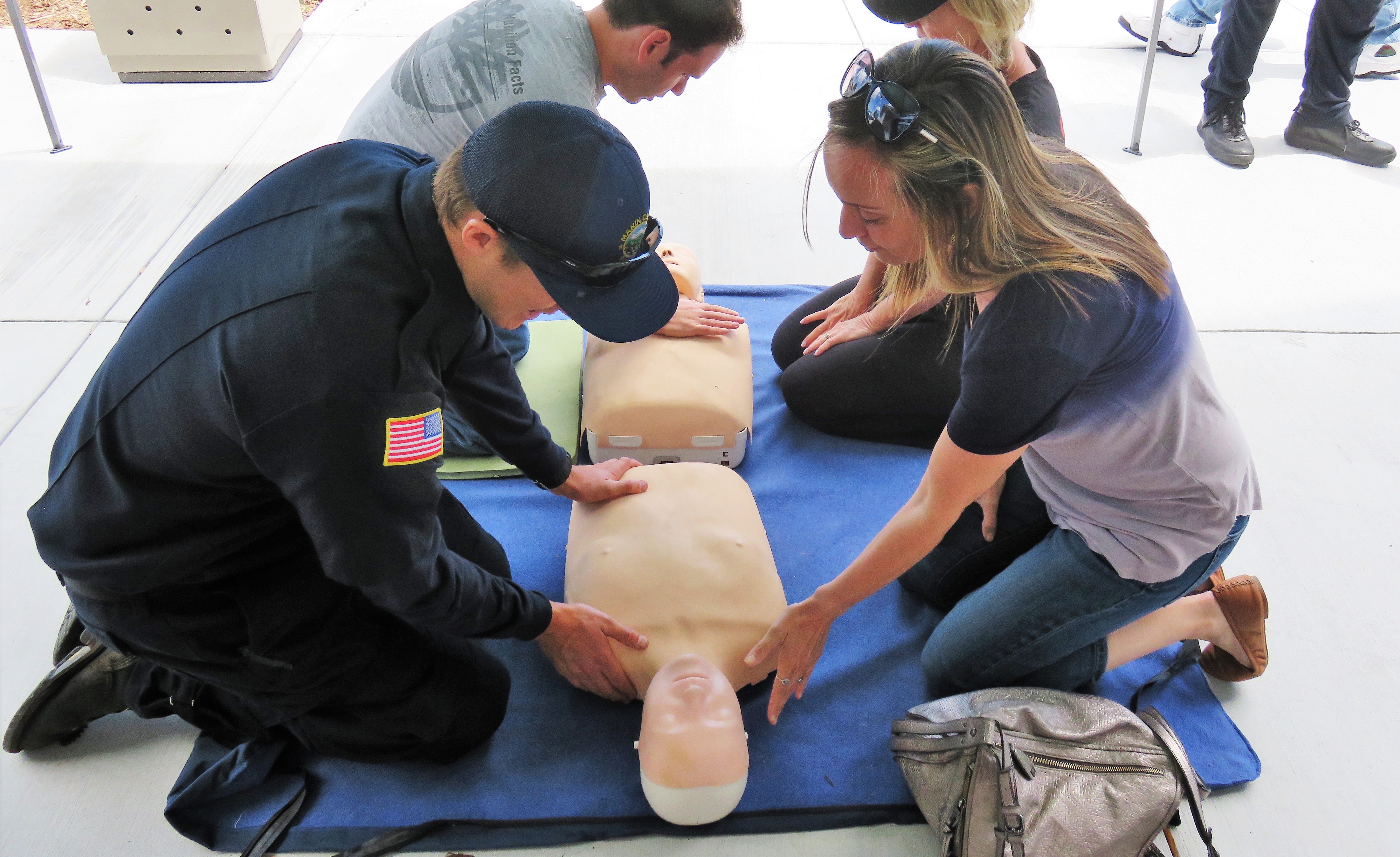 A male firefighter (left) teaches a woman (right) how to do CPR on a rubber dummy on a mat.