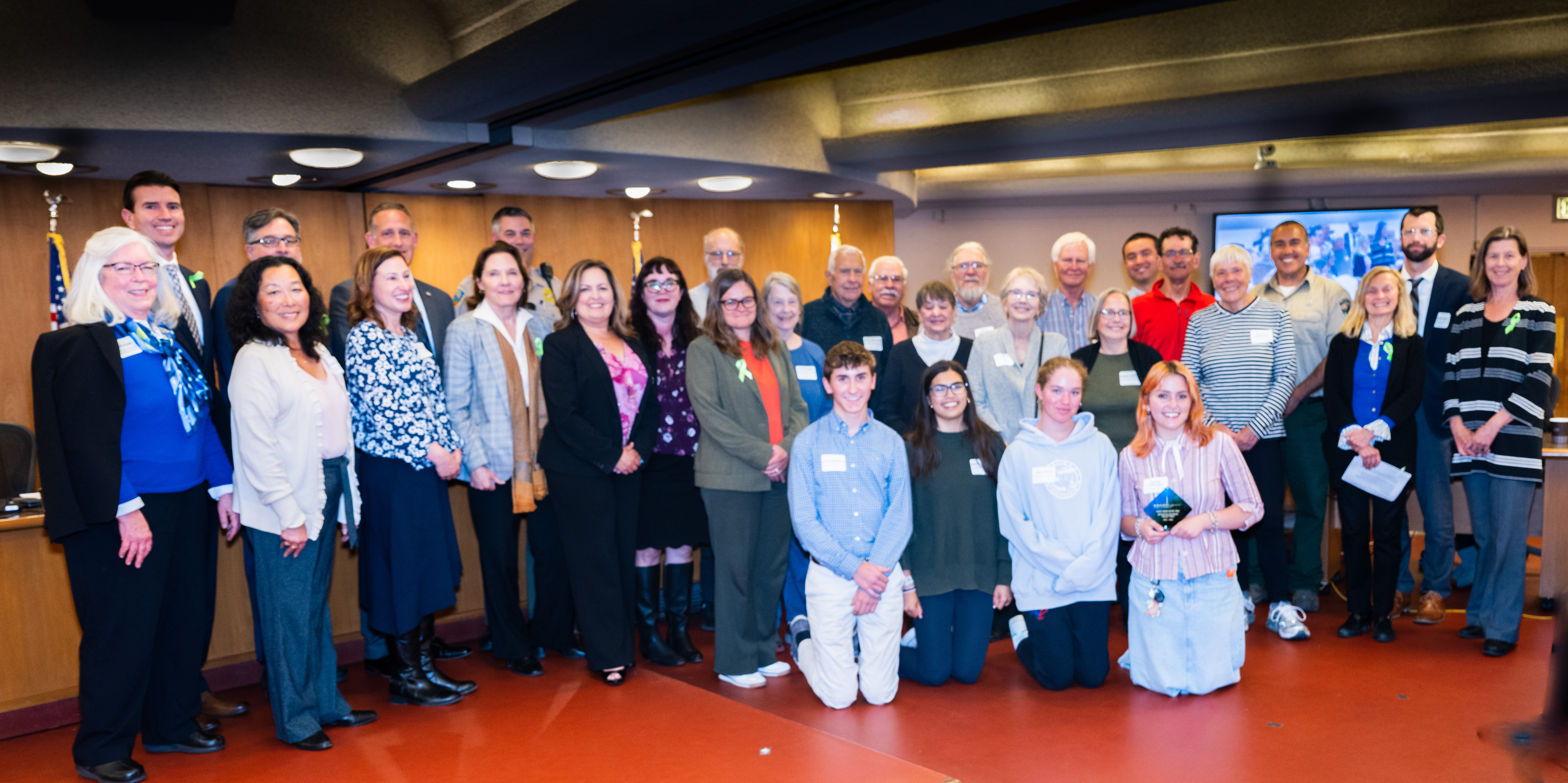 A group of about 25 volunteers smile and pose after being recognized by the Supervisors.