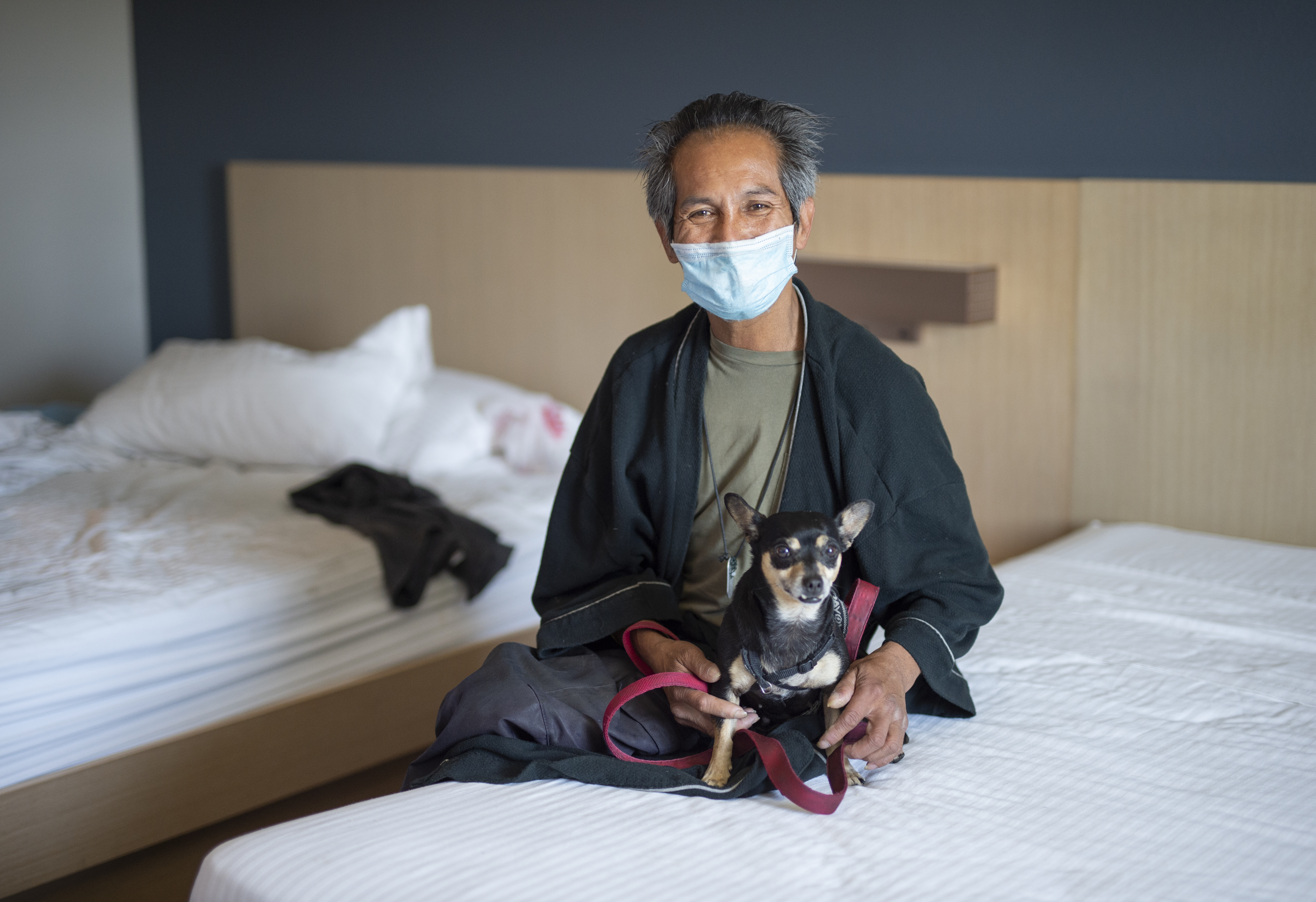 A man with a protective mask and a small dog sit on a bed.