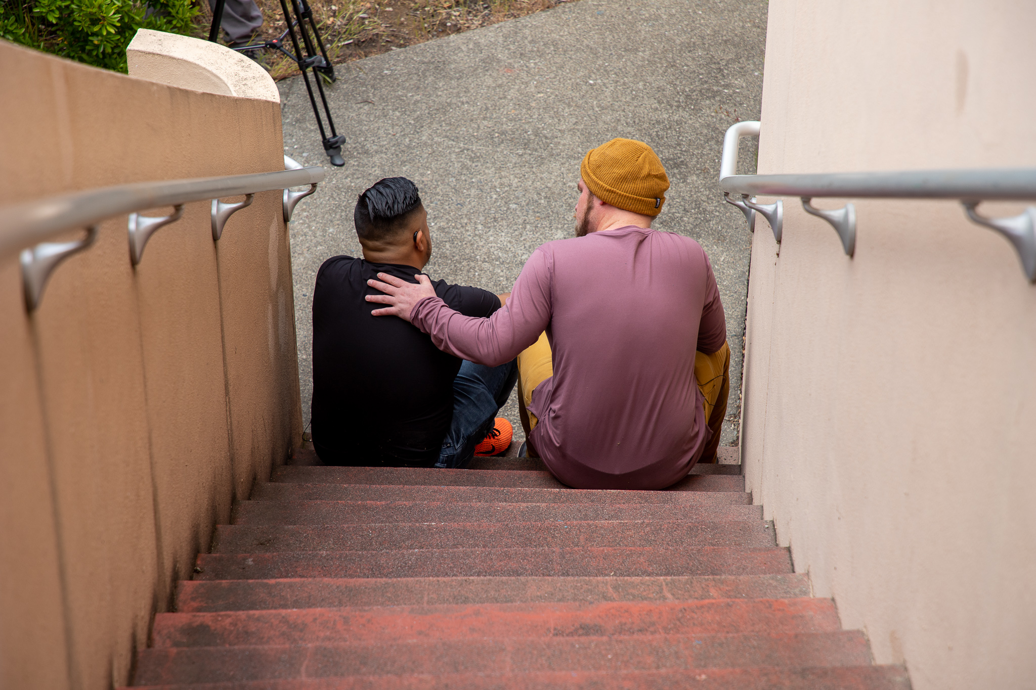 Two young men sit next to each other with the hand of one on the back of the other in support.