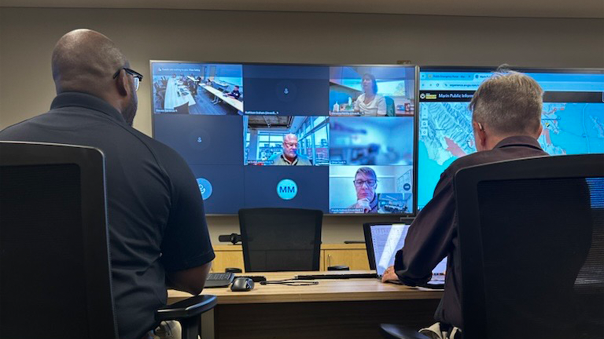 View of Emergency Operations Center conference room: Two men are seated, facing multiple monitors displaying a map and people participating in a virtual meeting. 