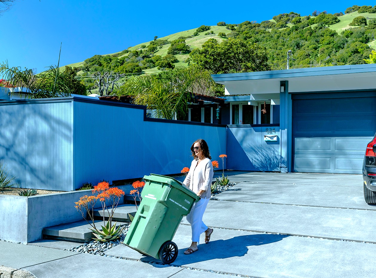 A woman brings a recycling bin out to a home's curb.