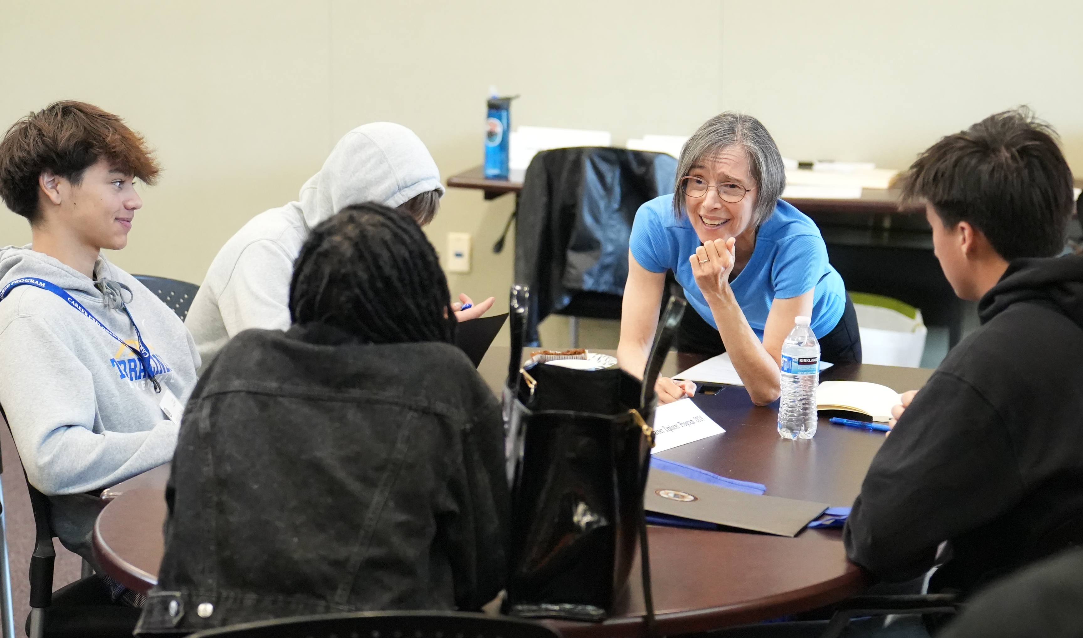 A woman leans over a table and speaks to several teenagers seated at the table. 