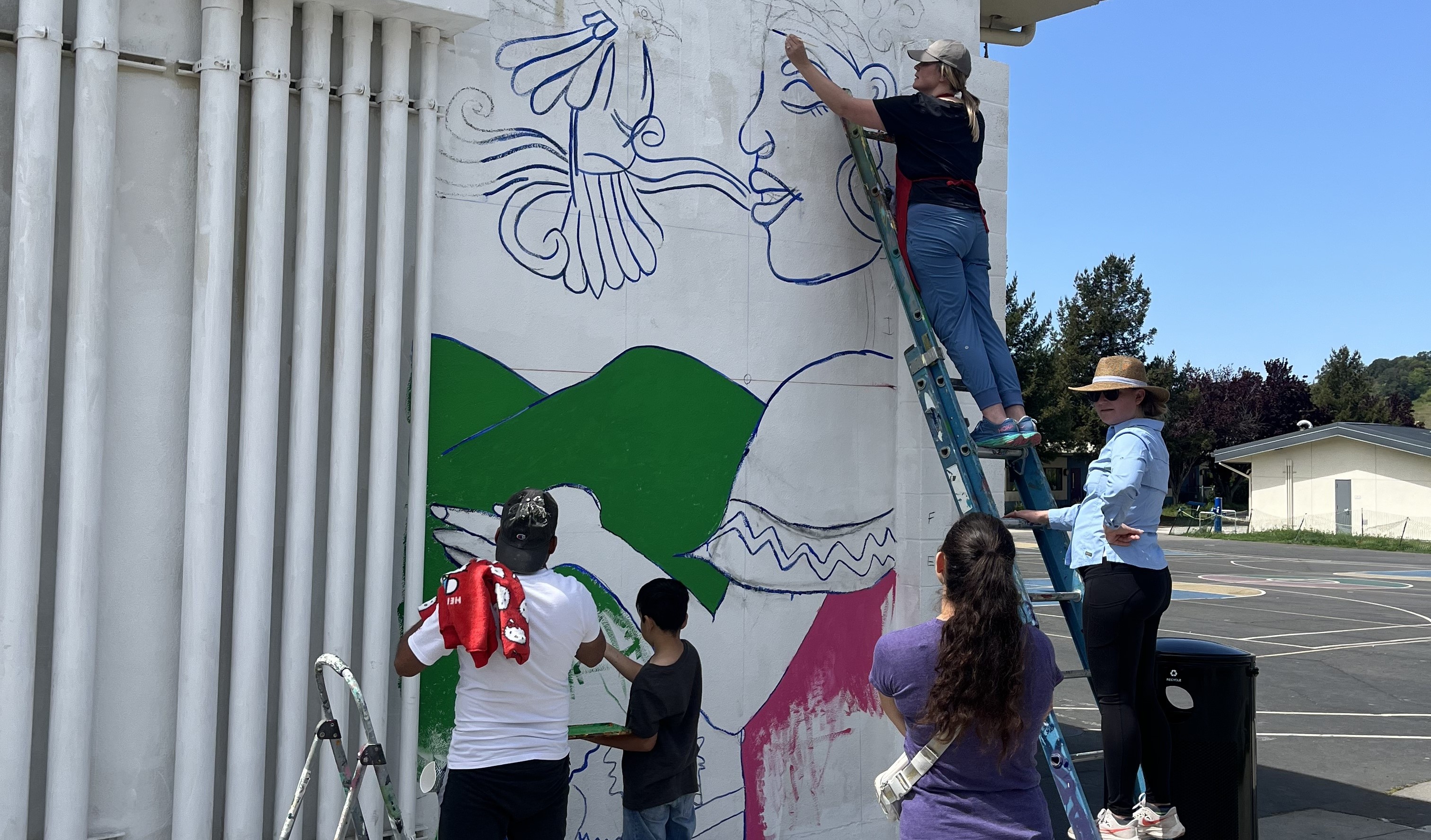 Community members paint a mural together on a school wall in a sunny schoolyard.