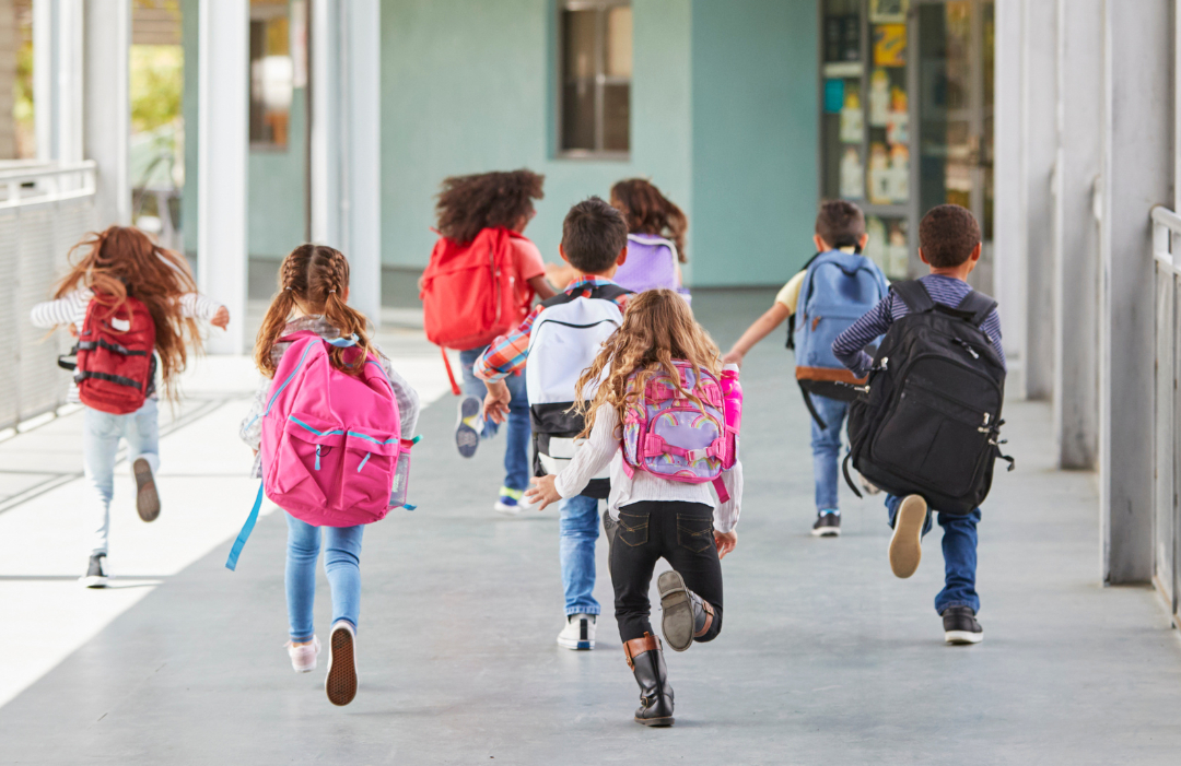 A group of young kids run down an outdoor hallway at a school.