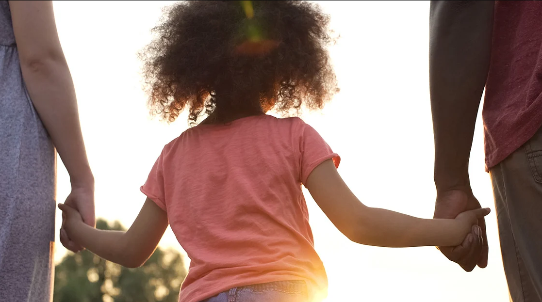 Child holding hands with two adults at sunset.
