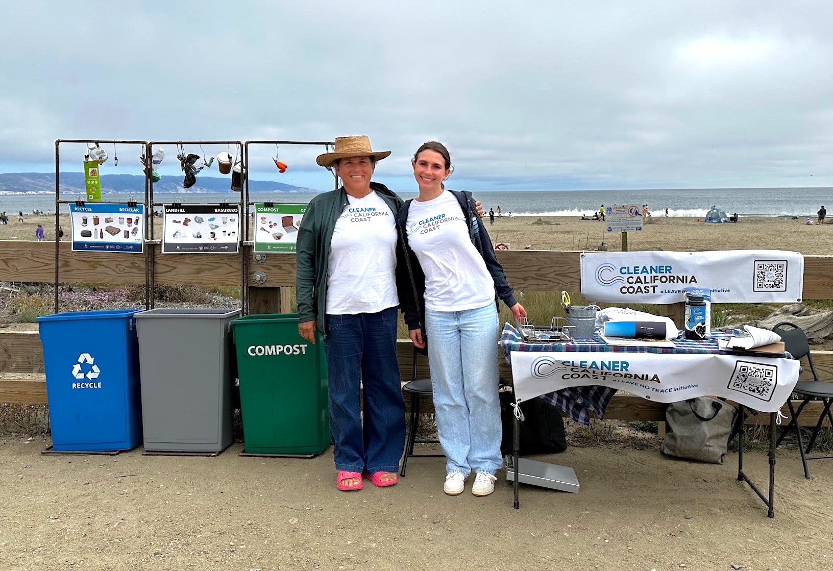 Two women pose at a Cleaner California Coast display at a beach event.