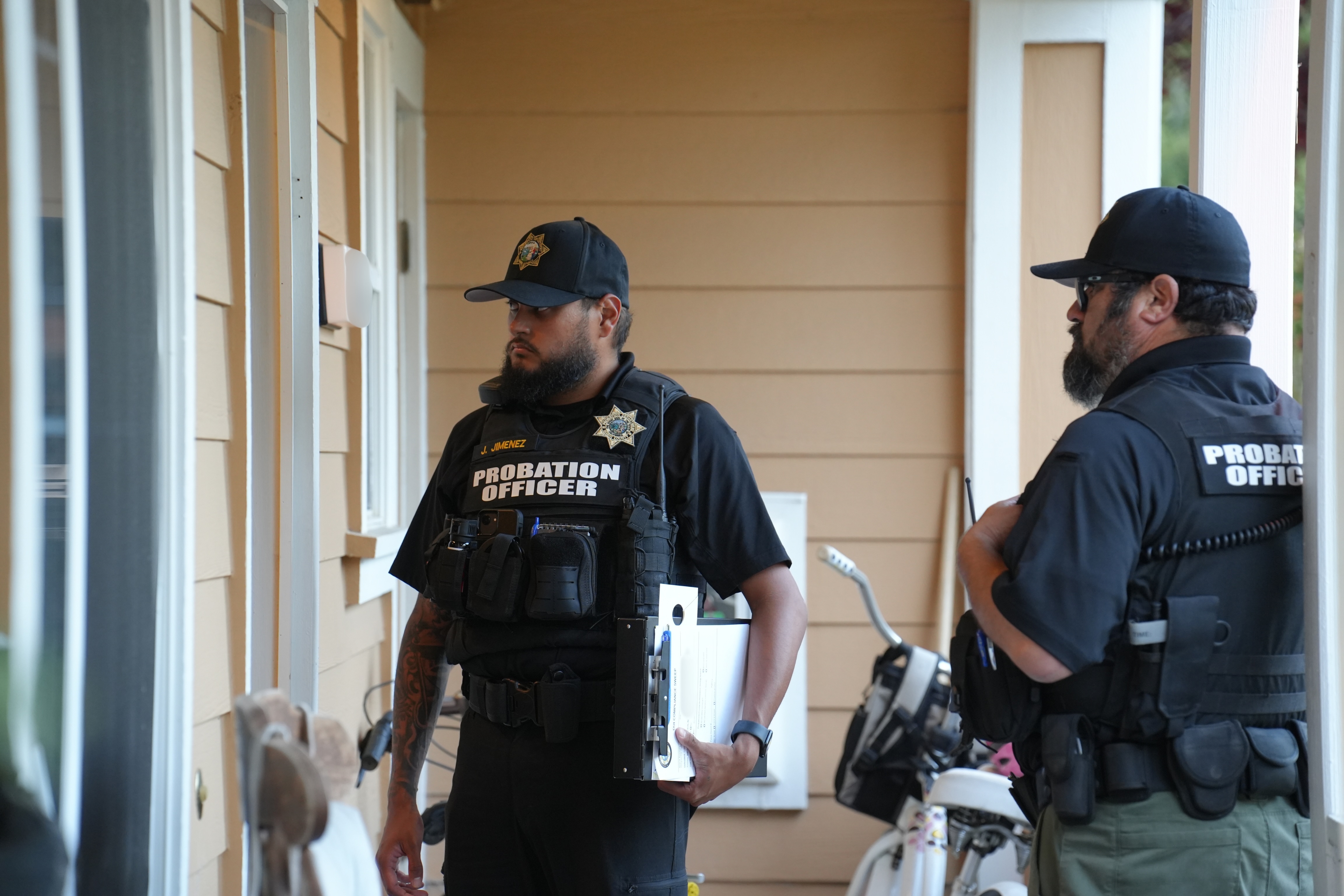 Two probation officers stand outside a residence waiting for someone to open a door.