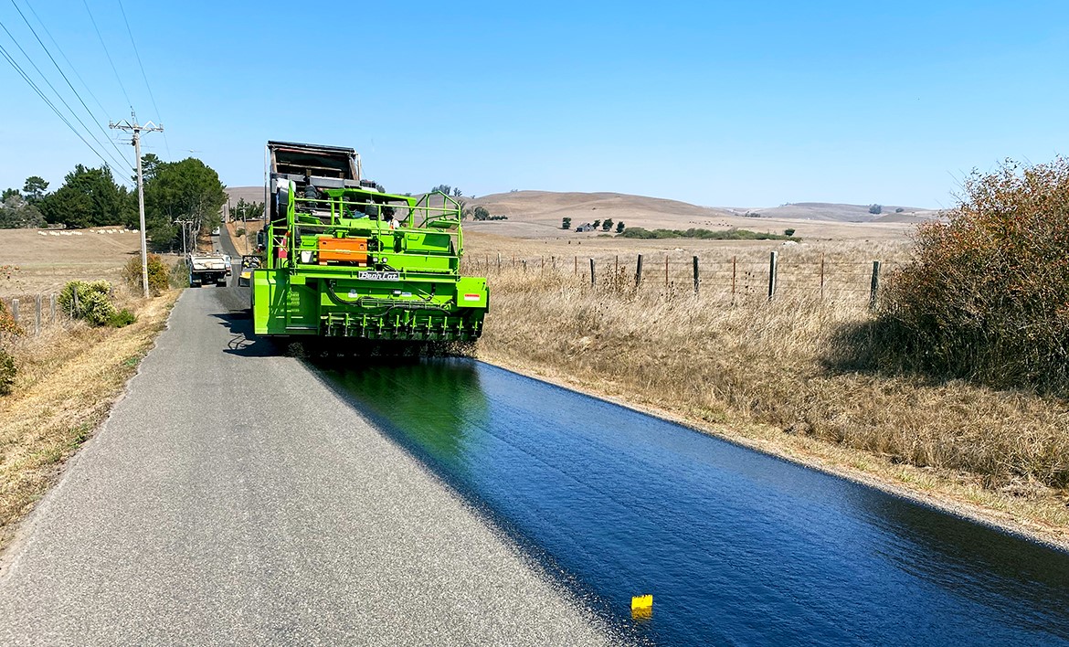 A large piece of heavy equipment is shown laying down fresh pavement on a rural road.
