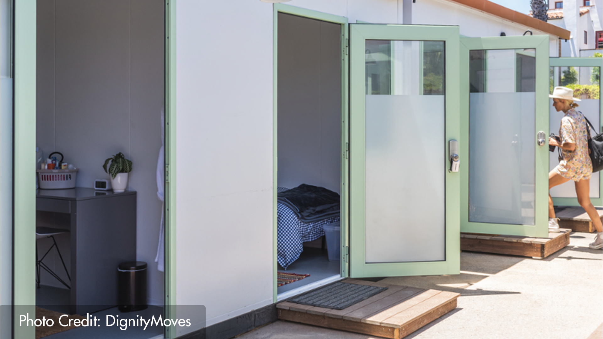 A representative tiny home community showing four units with doors open, and a woman walking into one unit.
