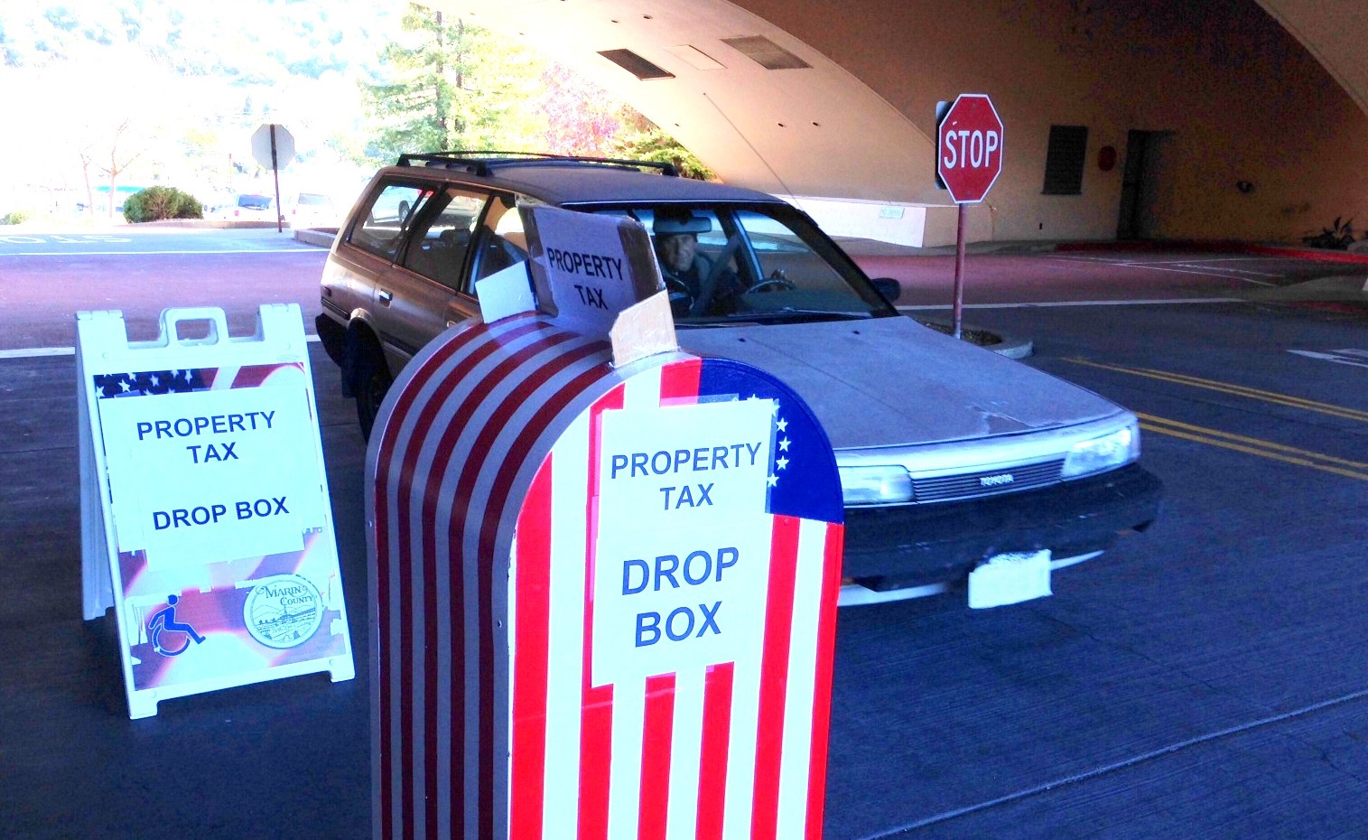 A driver in a car is stopped in front of a property tax dropbox outside the Civic Center. 