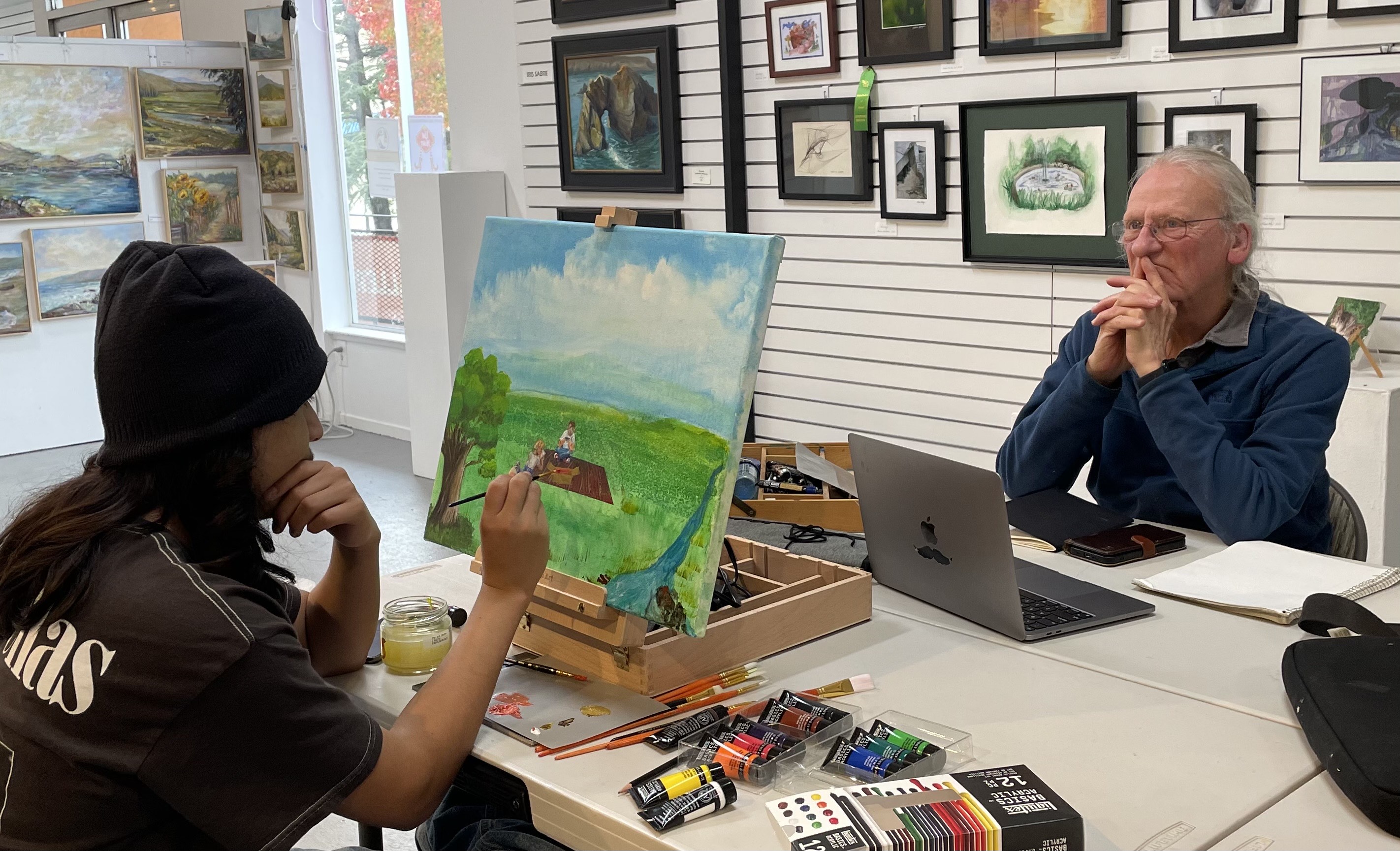 An older man sits as a desk as a younger boy creates the main's portrait.