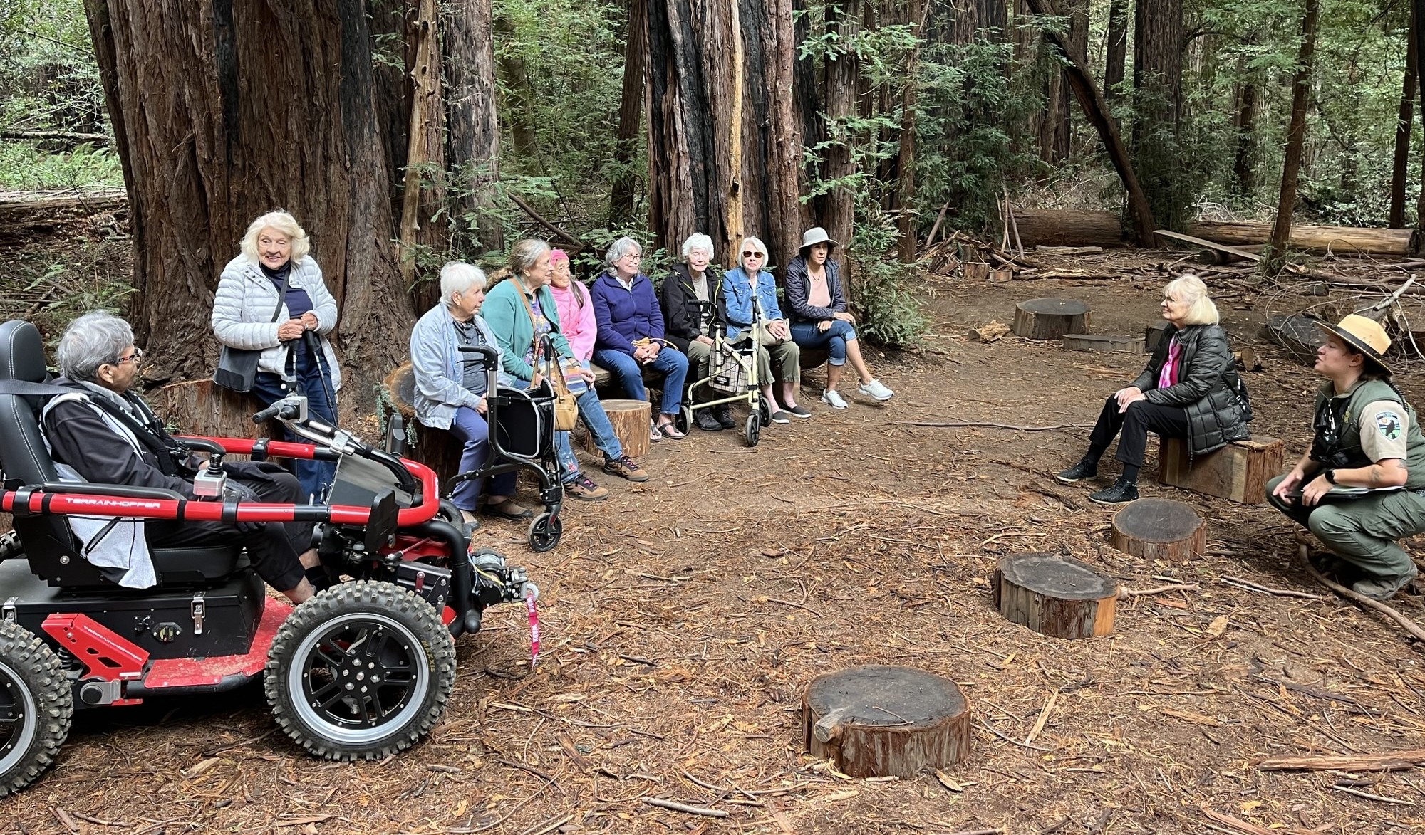 A small group of seniors listen to a parks ranger in a redwood forest grove.