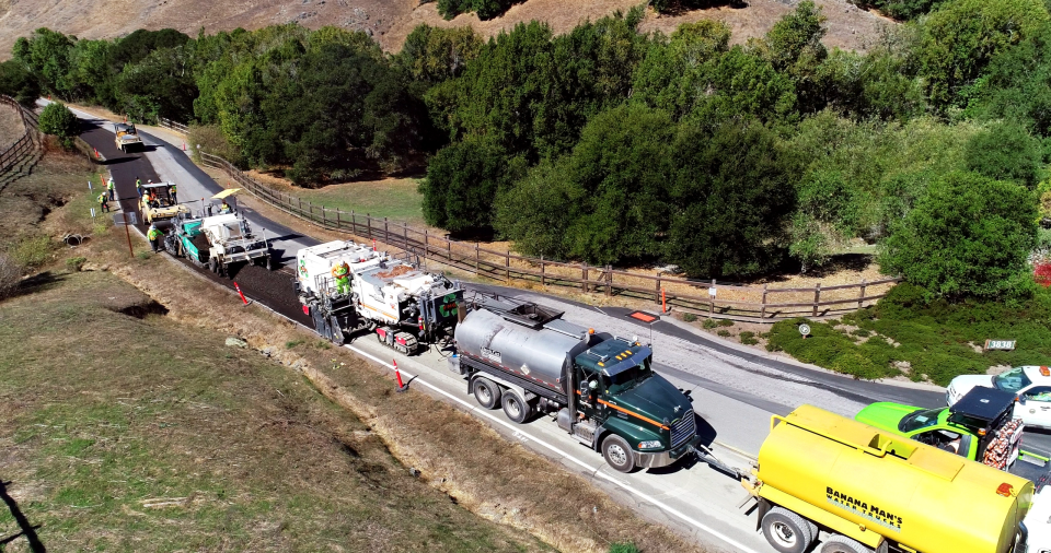 Several trucks are lined up on a road that it being repaved.