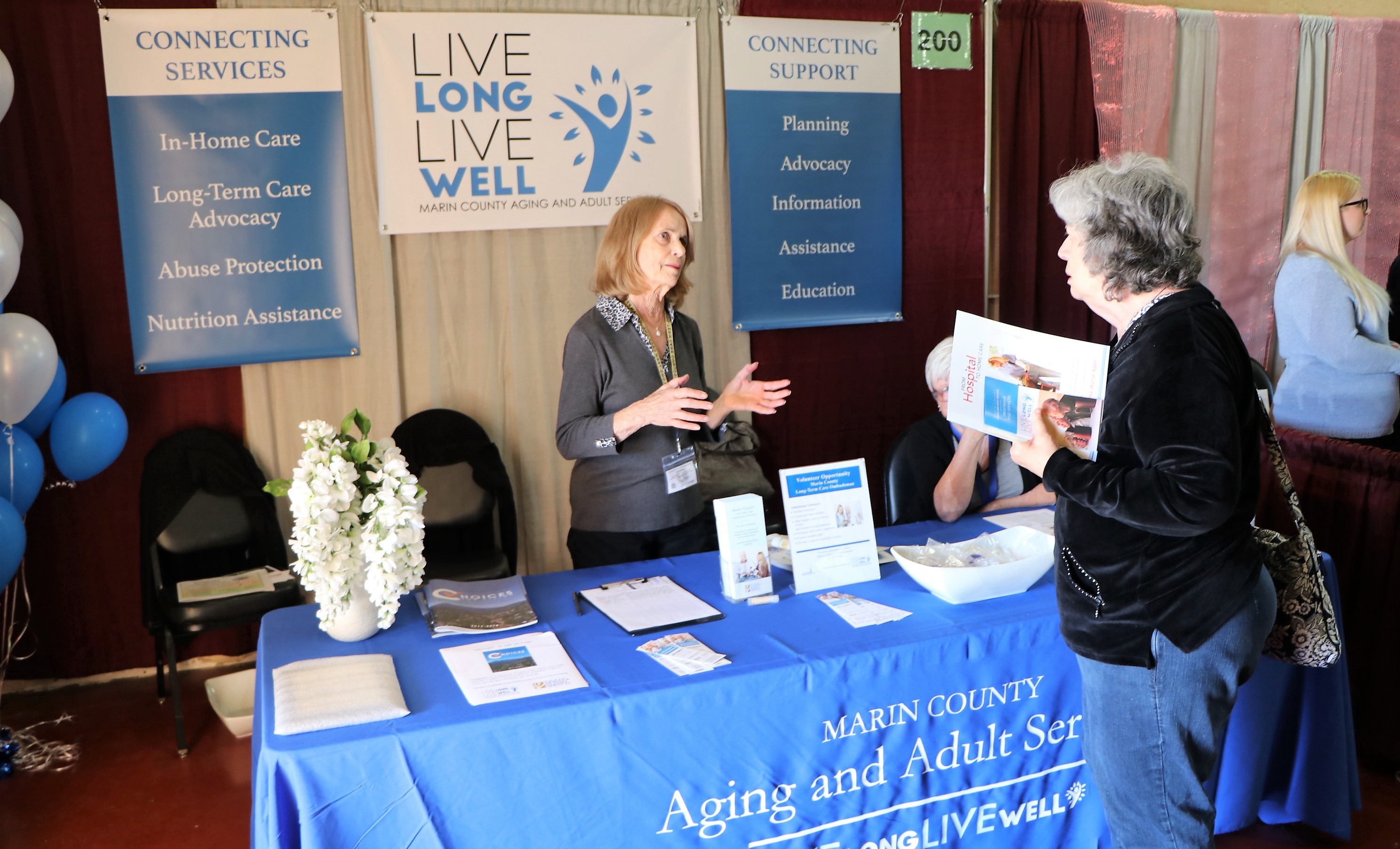 A woman at the Aging and Adult Services booth speaks to an elderly female attendee at a senior fair.