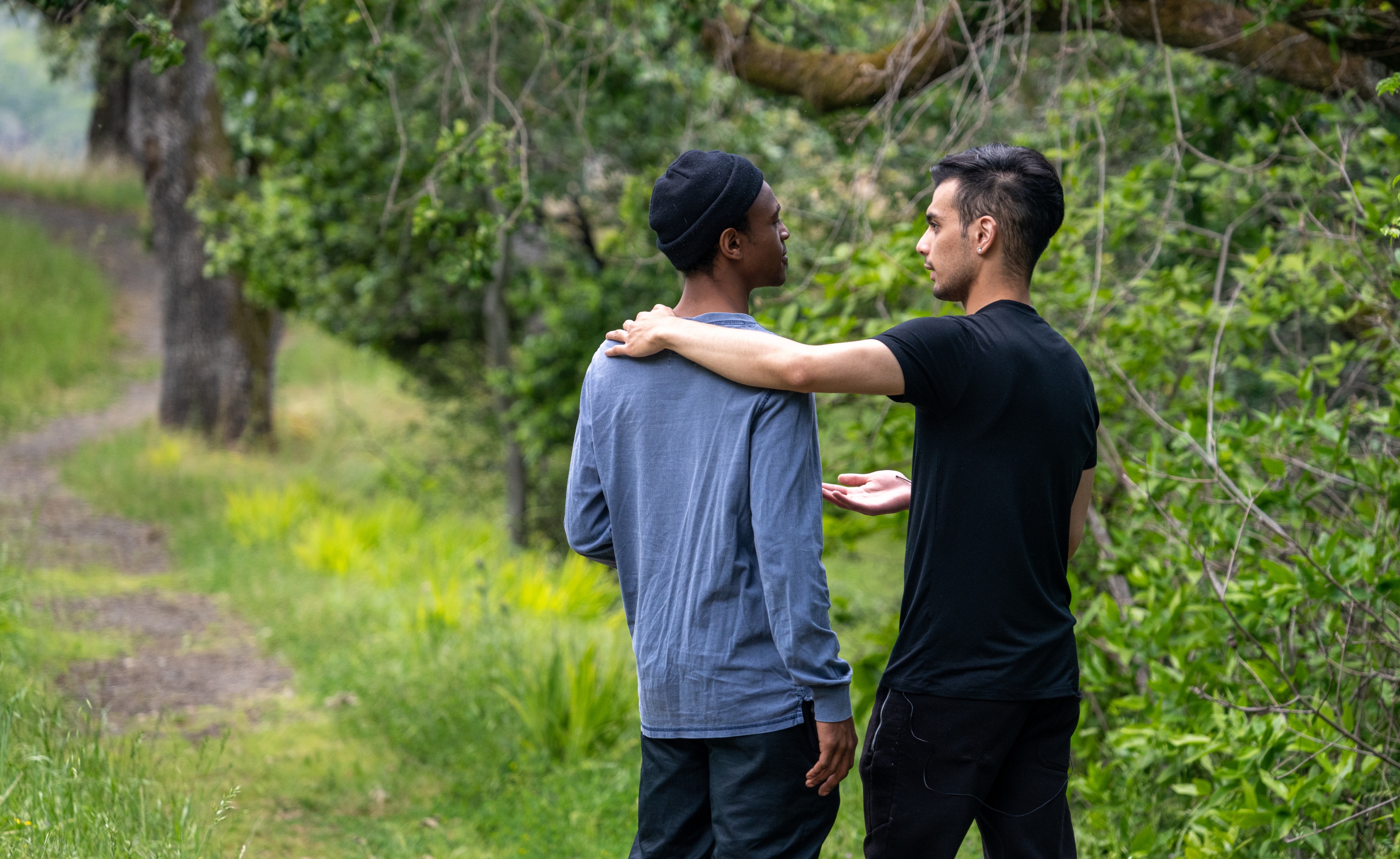 Two men stand in a park, with one man putting his arm around the shoulder of the other in a show of support.
