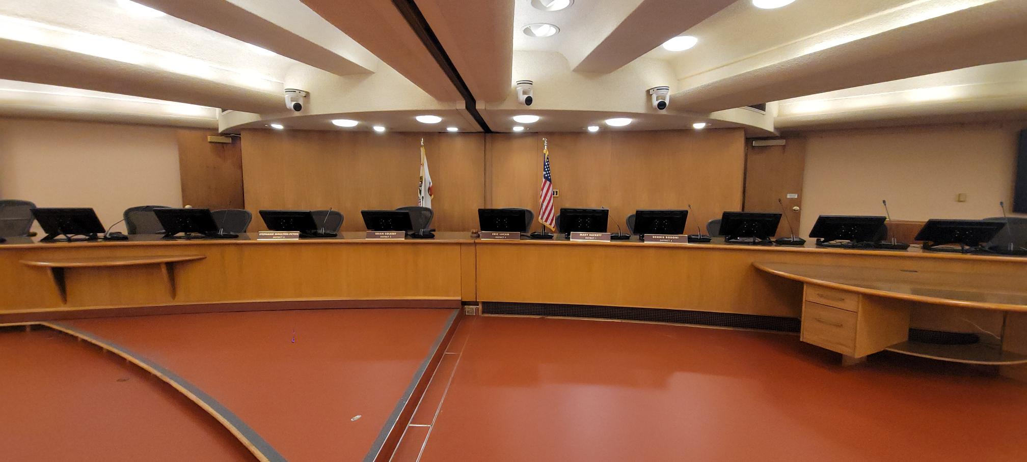 The BOS Chamber wooden dais with multiple seats, microphones, and monitors, set beneath recessed lighting and security cameras, with the U.S. and California flags displayed at the center.