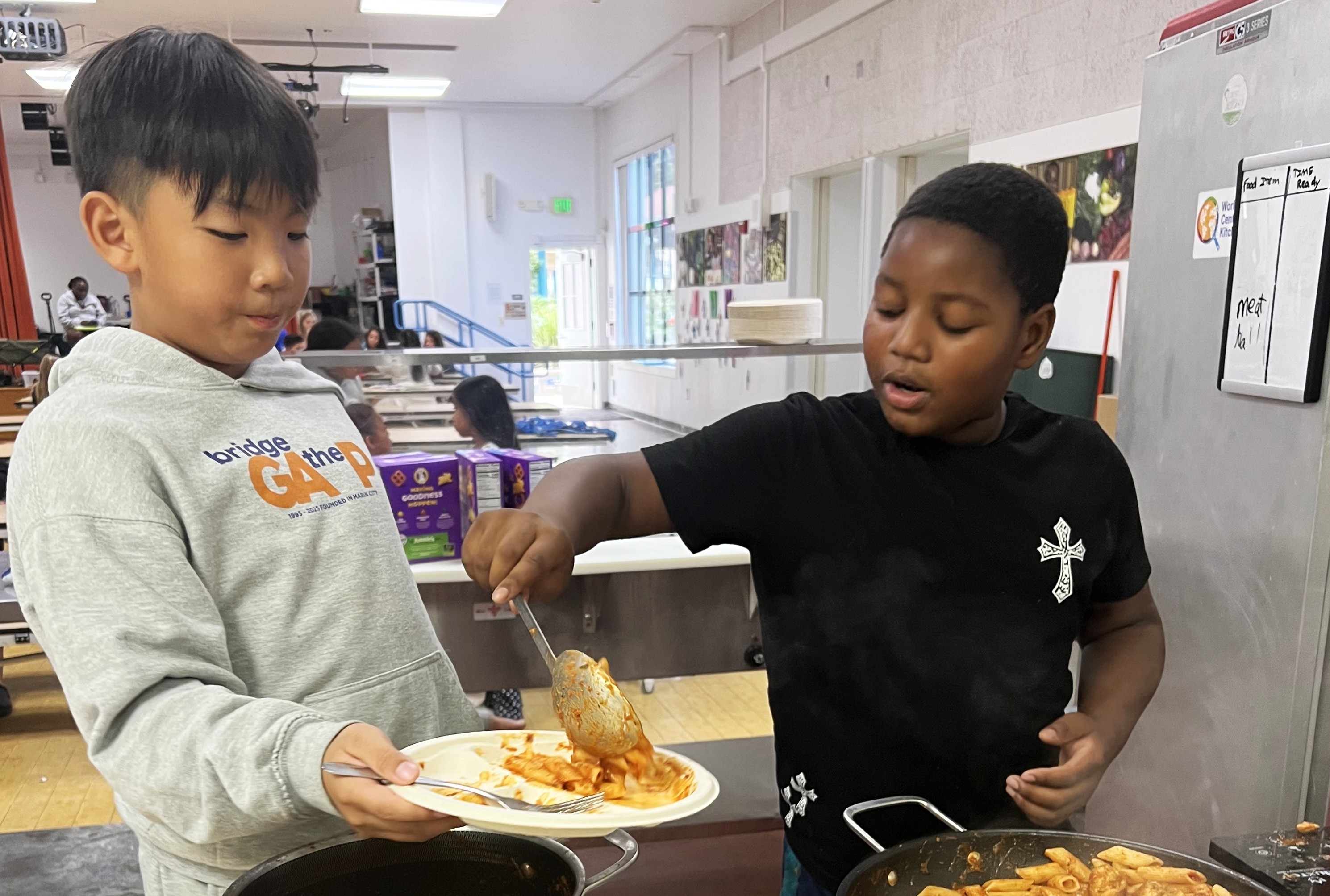 One young boy serves food to another boy holding a plate.