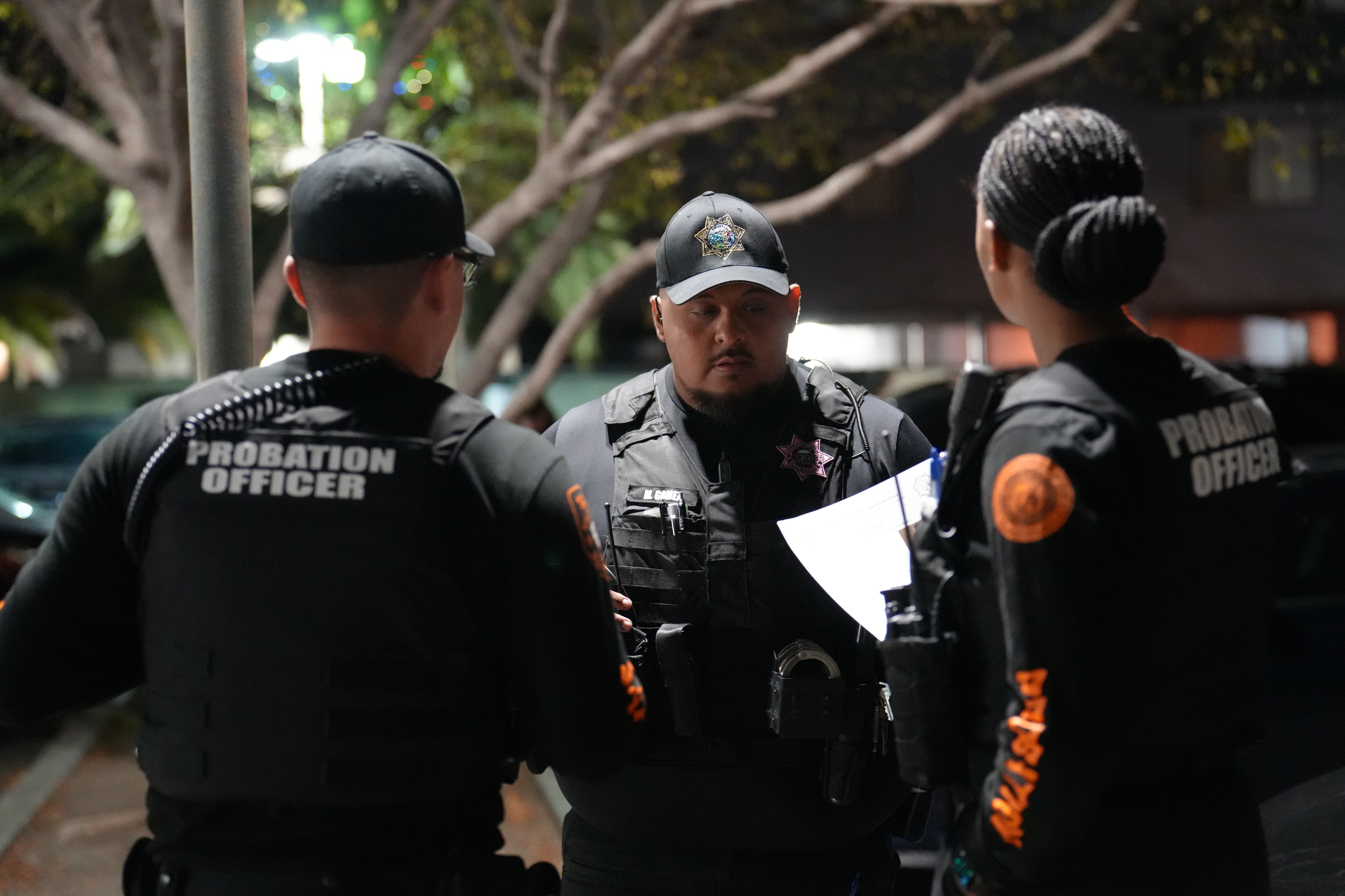 Three probation officers in tactical gear stand outdoors at night reviewing notes during a briefing.