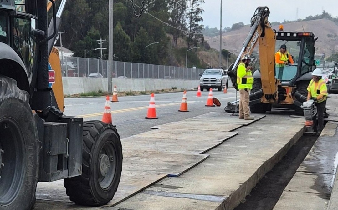 Workers wearing hardhats operate heavy equiptment on a roadway project.