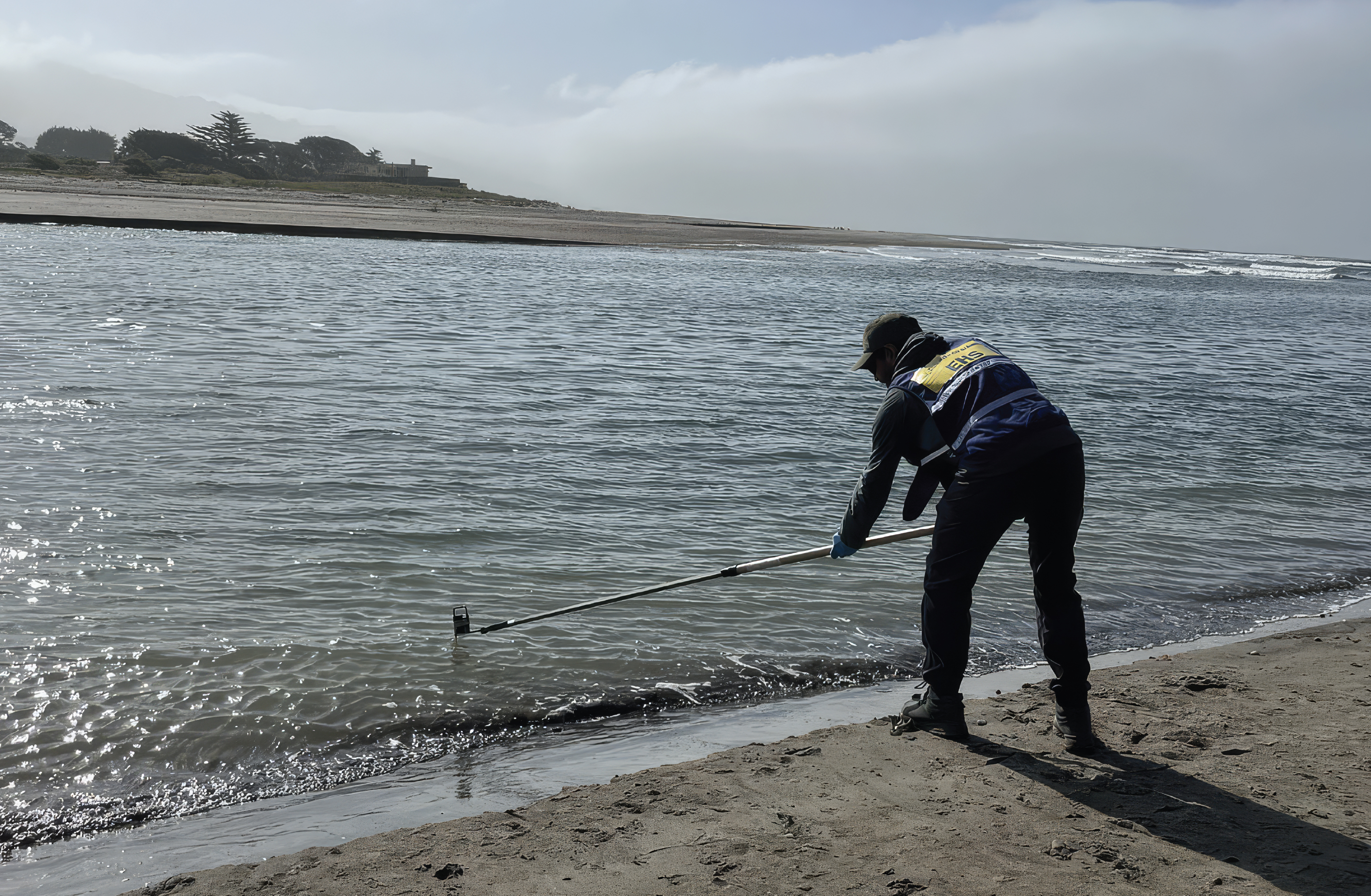 A technician sands on a beach and reaches a long pole into bay waters to retieve a test sample
