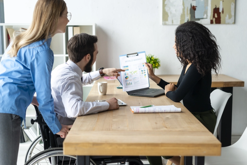 Three people sit at a table in an office and point to a paper on a clipboard.