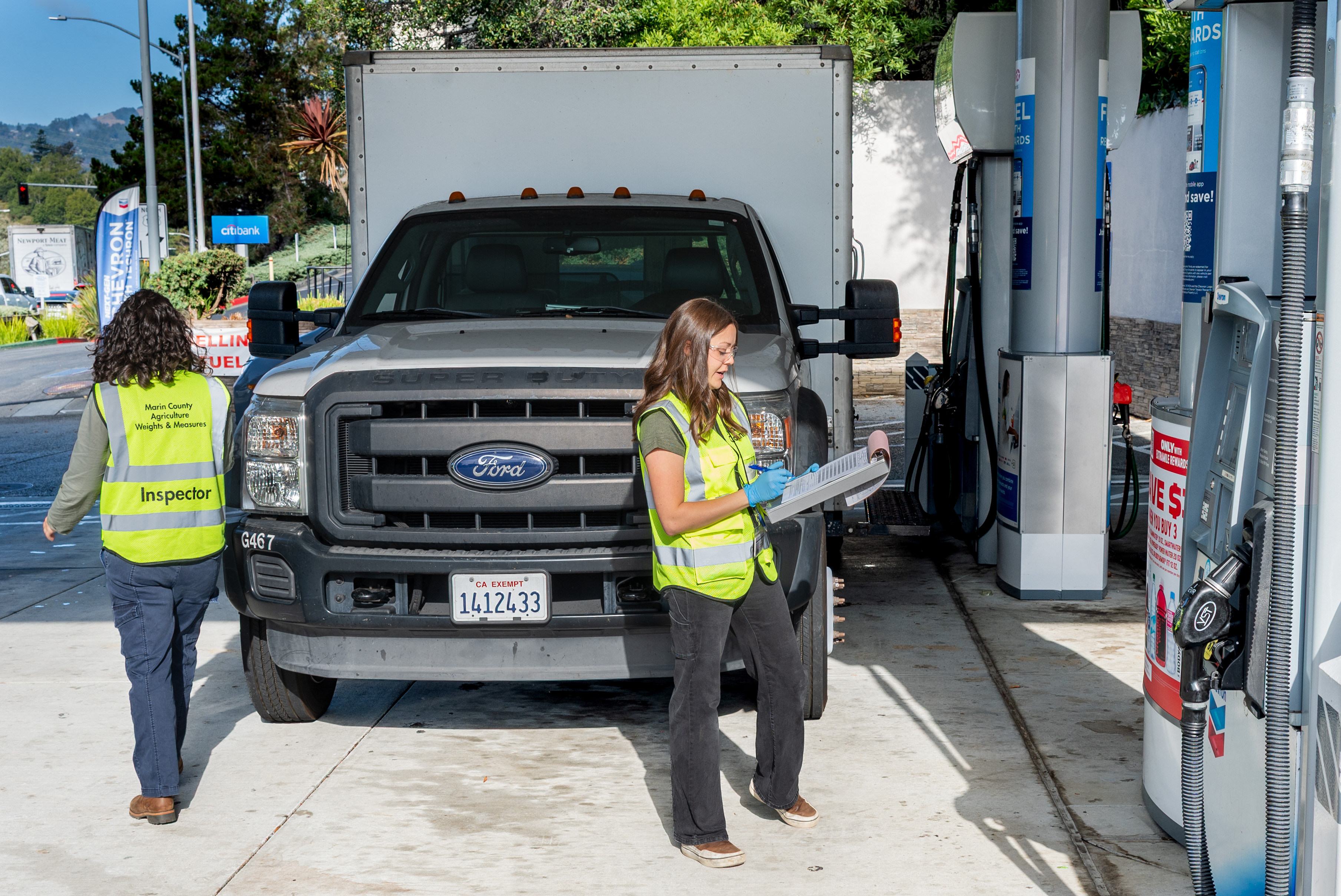 Two female inspectors stand next to a truck and test gasoline pumps at a station.