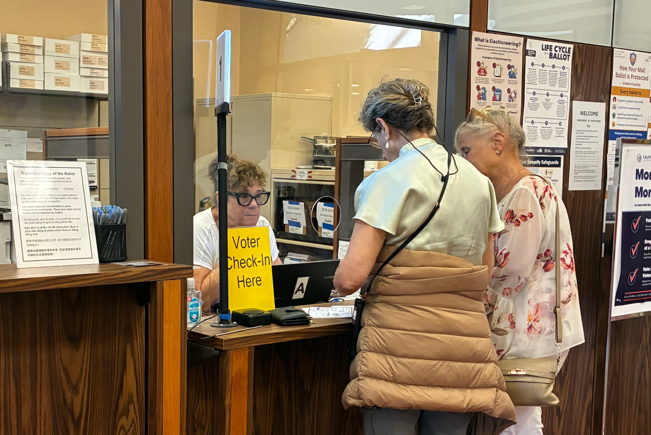 Two women stand at the front desk of the Elections Department and speak to a staff remember.