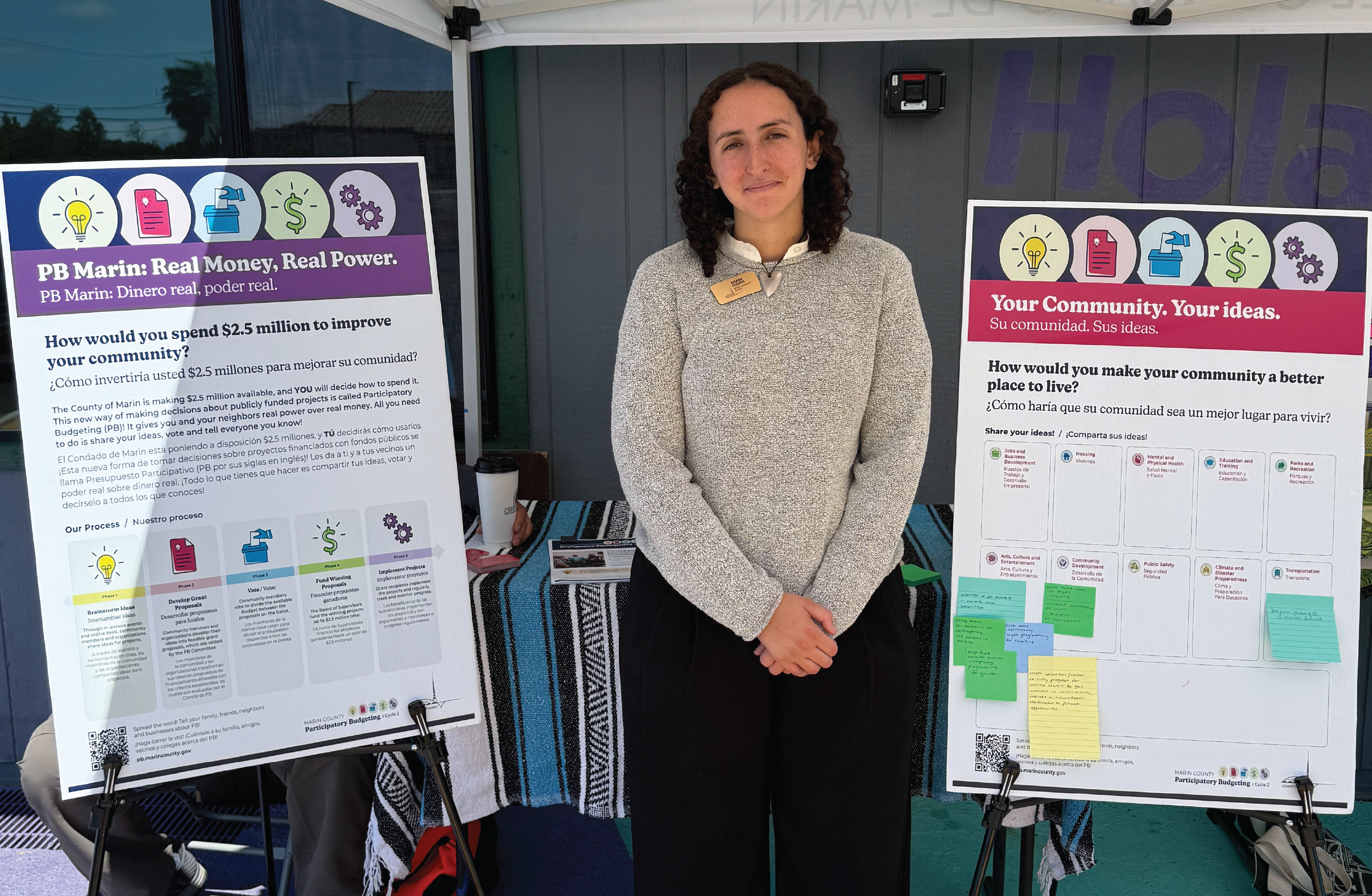 Participatory Budgeting staff member stands between two bilingual community engagement boards inviting residents to share ideas for improving their community.