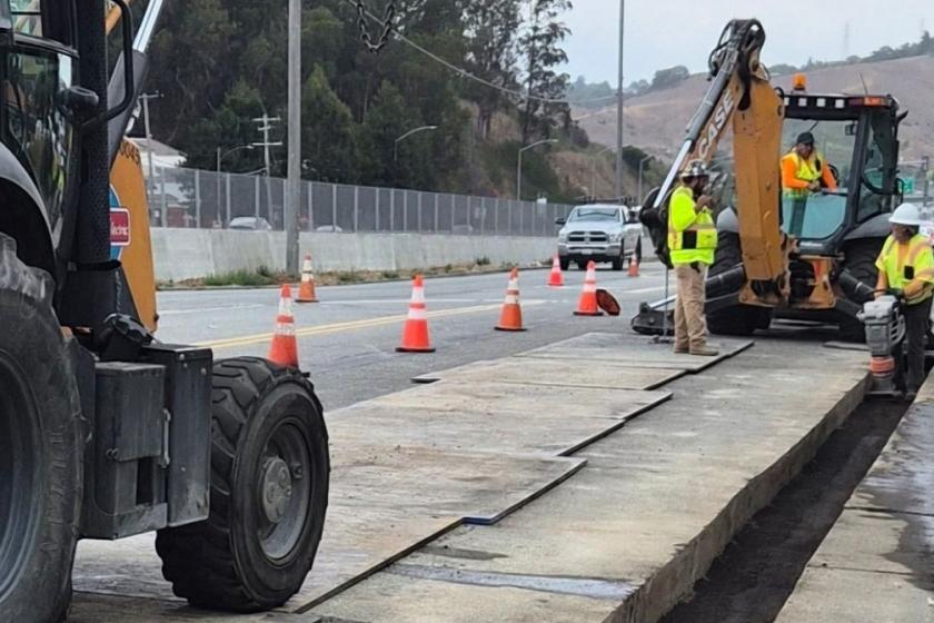 Workers wearing hardhats operate heavy equiptment on a roadway project.