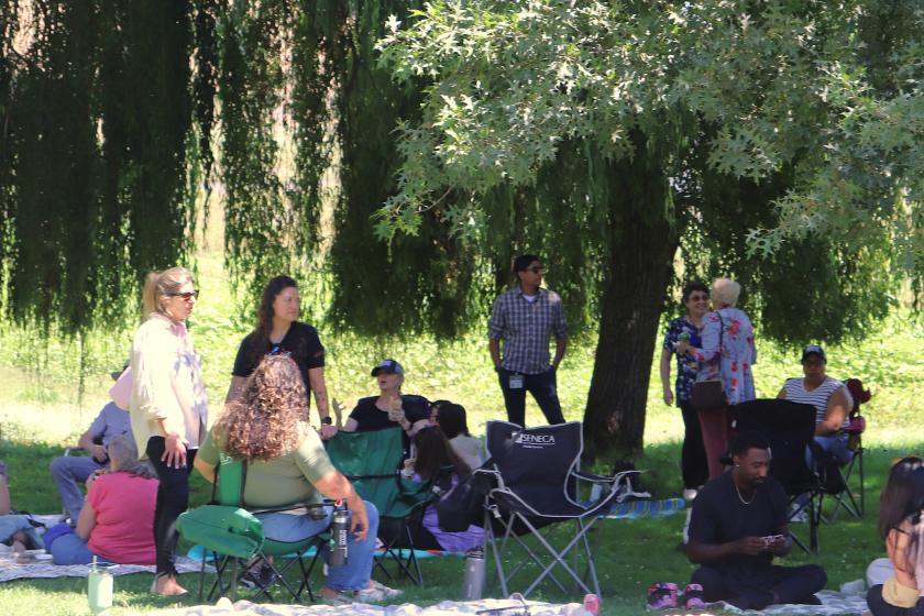 About a dozen people sit in the shade of a tree at a park.