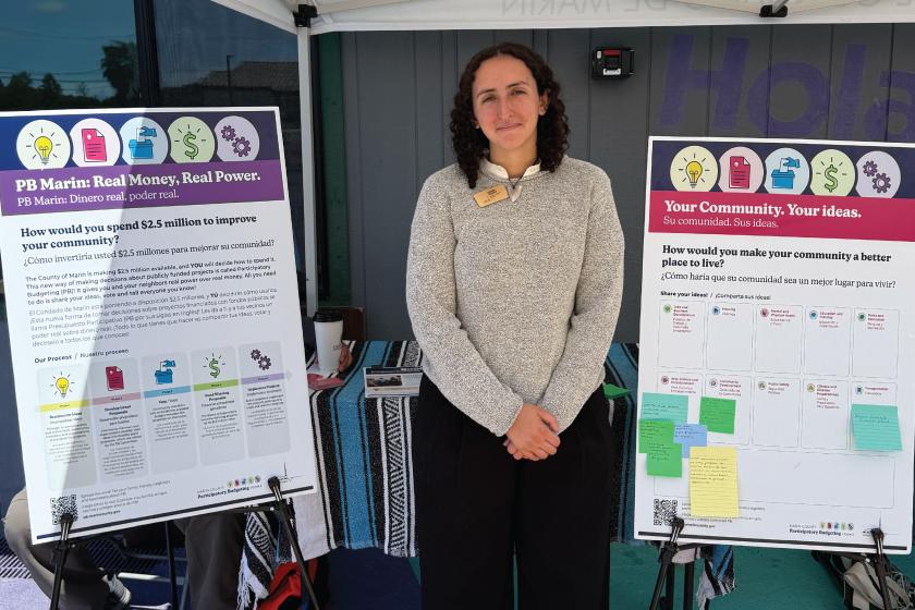 Participatory Budgeting staff member stands between two bilingual community engagement boards inviting residents to share ideas for improving their community.