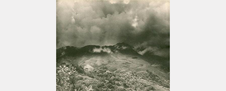 Clouds of smoke over a mountain, with low brush in the foreground. Black and white photo.