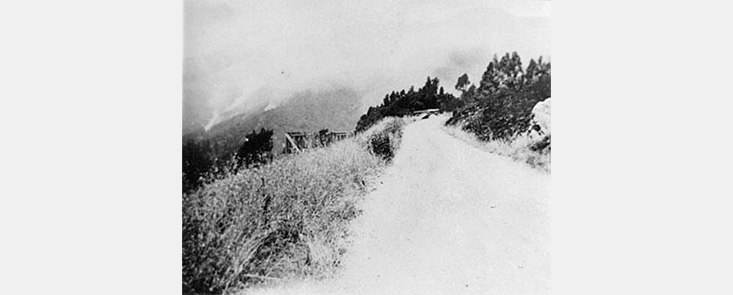 A road cuts through a hillside, with smoke in background. Black and white photo.