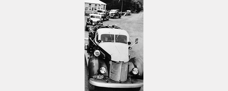 A line of 5 1950s cars along a road. Black and white photo.