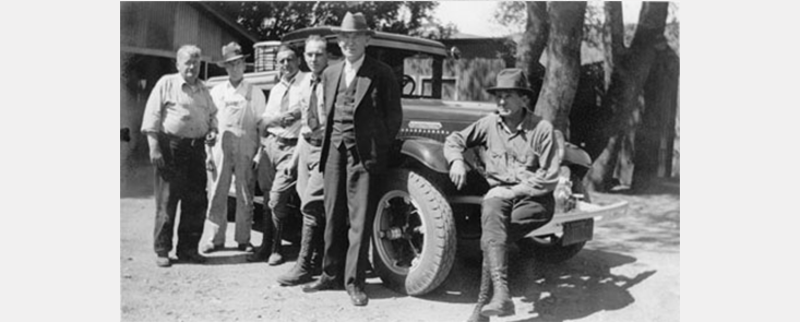 6 men standing around a car. Edwin Gardner stands in front wearing a suit. Black and white photo.