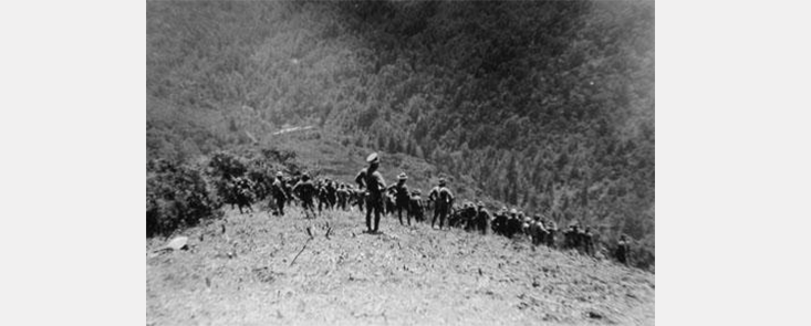 A large group of firefighters standing on a hill, looking out at smoke. Black and white photo.