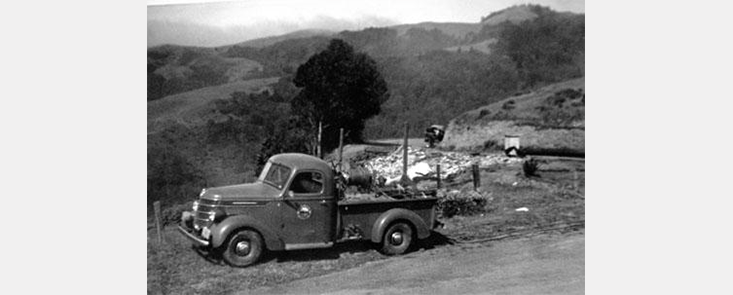 A car on the side of a road with hills in the background. Black and white photo.