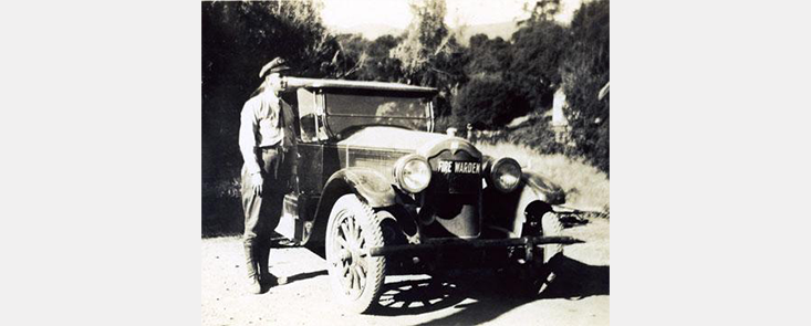 A man in fire uniform standing next to a car. The car obscures the background. Black and white photo.