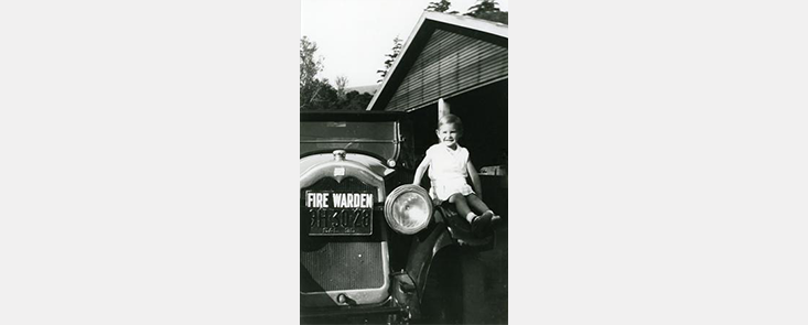 A young girl sitting on the hood of a car outside a building. Black and white photo.