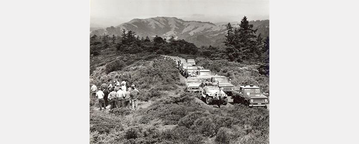 A large group of men standing next to a line of cars, mountains and trees in the background. Black and white photo.