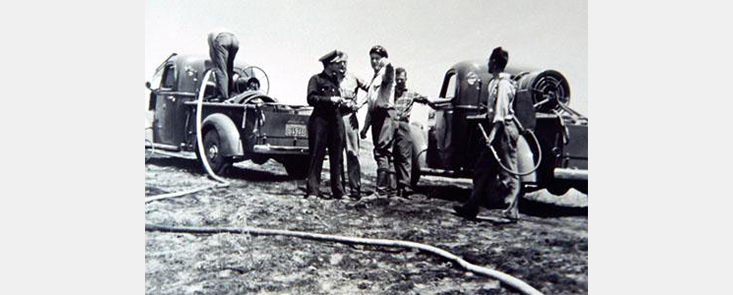 6 men attaching hoses to 2 fire trucks. The background is smokey. Black and white photo.